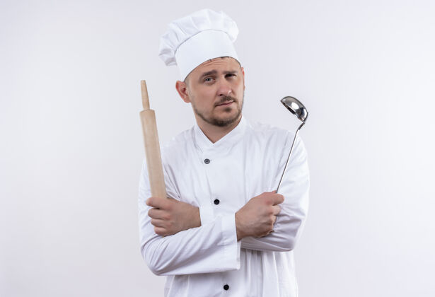 卷自信的年轻帅哥厨师身着厨师制服 拿着勺子和擀面杖 站在隔离的白色墙壁上 留着复制空间别针自信厨师