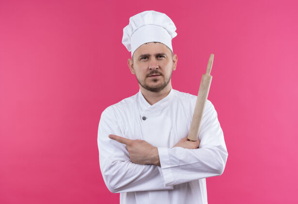 滚自信的年轻帅哥厨师穿着厨师制服拿着擀面杖指着粉色墙上孤立的一面指着帅气别针