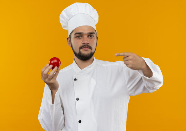 厨师身着厨师制服的自信年轻男厨师手拿并指着隔离在橙色墙上的西红柿拿着自信制服