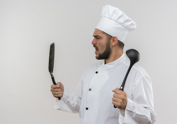 烹饪愤怒的年轻男厨师穿着厨师制服 拿着煎锅和勺子看着隔离在白墙上的锅年轻制服男