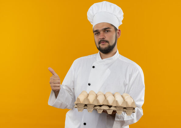 纸箱自信的年轻男性厨师身着厨师制服 手拿一盒鸡蛋 在橙色的墙壁上孤立地竖起大拇指 留有复印空间显示男性持有