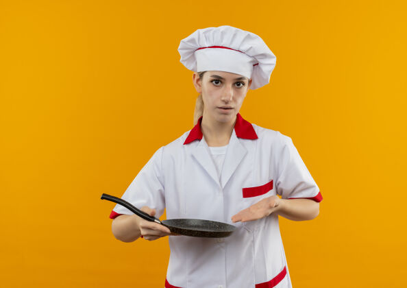 平底锅年轻漂亮的厨师穿着厨师制服 拿着并指着隔离在橙色墙上的煎锅年轻制服拿着