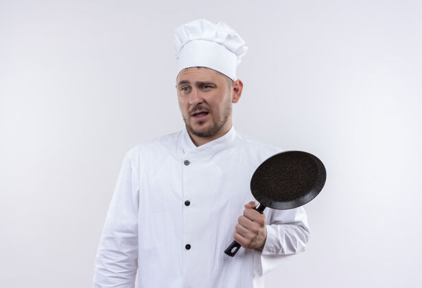 壁板迷茫的年轻帅哥厨师穿着厨师制服拿着煎锅看着隔离在白墙上的一面困惑平底锅烹饪
