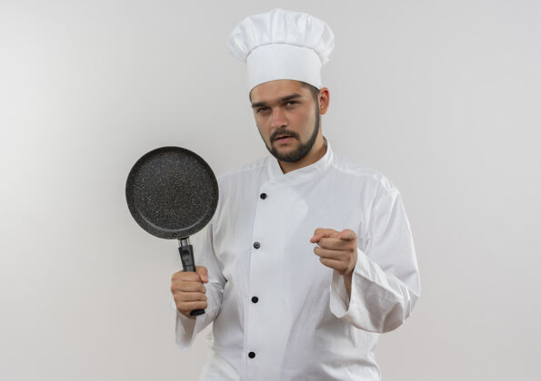 指着自信的年轻男厨师身着厨师制服 手拿煎锅 指着隔离在白墙上的墙拿着烹饪平底锅