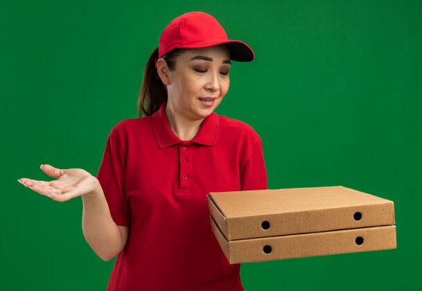 年轻年轻的送货员身穿红色制服 戴着帽子 手里拿着披萨盒 站在绿色的墙上 困惑地伸着胳膊帽子胳膊拿着