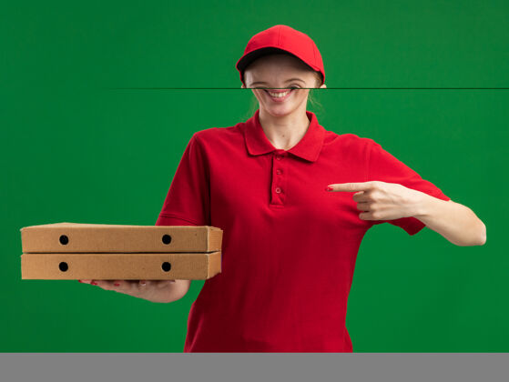 年轻身穿红色制服 头戴鸭舌帽 手持披萨盒 食指指着披萨盒的年轻送货女孩站在绿色墙壁上自信地微笑着盒子披萨手指