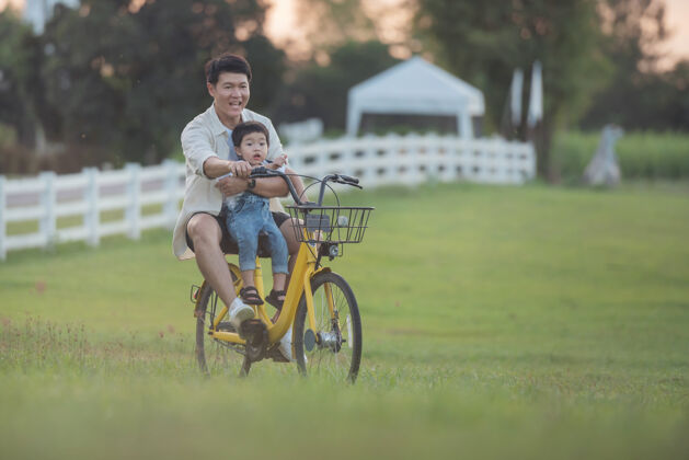 人一对骑着自行车的快乐的年轻父亲和儿子的画像日落时分 父亲和儿子在公园里玩耍人们在球场上玩耍友好家庭和暑假的概念亚洲人活动父亲