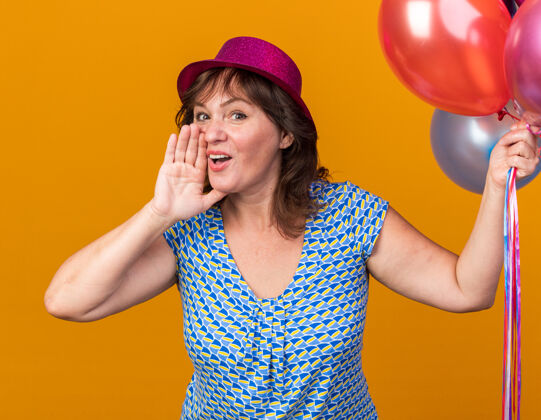 喊戴着派对帽的中年妇女手持五颜六色的气球 用手在嘴边喊着快乐快乐的生日派对站在橘色的墙上举行中间五颜六色