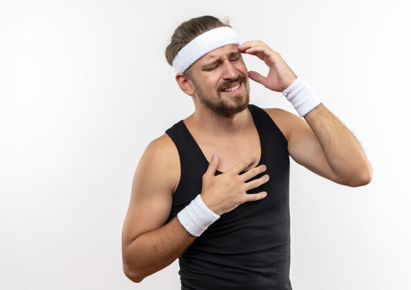 戴疼痛的年轻英俊的运动型男子戴着头带和腕带 双手放在太阳穴和胸前 闭着眼睛 隔离在白色的墙壁上 留着复制空间疼手胸部