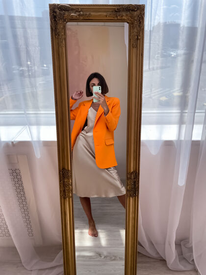 相机穿着浪漫的米色丝绸连衣裙和橙色外套的健康晒黑女人在家里对着镜子在电话里自拍时尚采取年轻