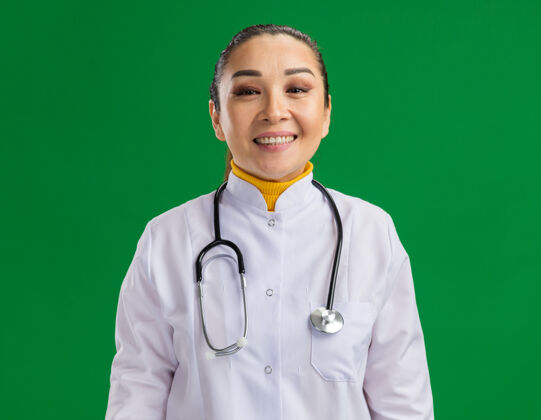 周围快乐的年轻女医生 穿着白药衣 脖子上戴着听诊器 站在绿色的墙上 微笑着 自信地站着站医生微笑