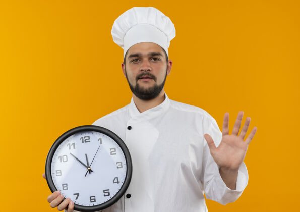 空自信的年轻男厨师 身着厨师制服 手拿时钟 在橙色的墙上孤零零地伸出空手年轻制服烹饪