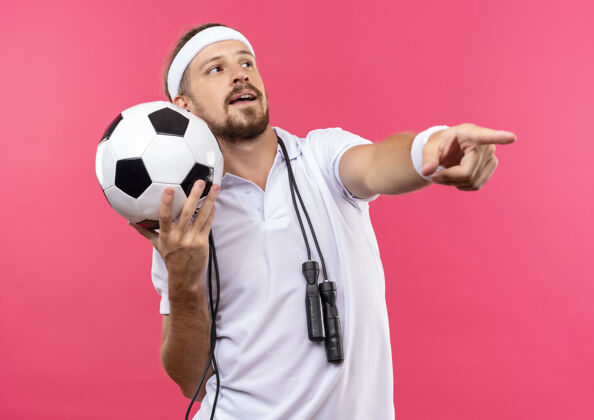 指向令人印象深刻的年轻英俊的运动男子戴着头带和腕带拿着足球看在一边 并直指与跳绳脖子隔离在粉红色的墙上男子绳子侧线