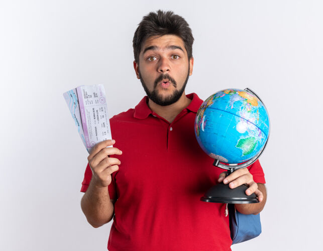 地球仪年轻的留着胡子的学生 穿着红色马球衫 背着背包 手里拿着机票 站着的地球仪看起来很惊讶地图地球仪年轻