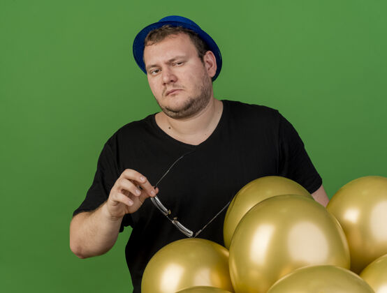 氦自信的成年斯拉夫男子手持眼镜 头戴蓝色派对帽 手持氦气球空间绿色气球
