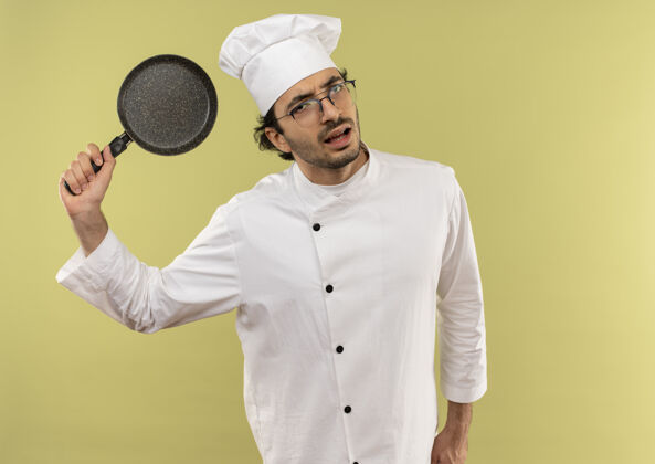 制服年轻的男厨师穿着厨师制服 戴着眼镜 在绿色的背景下举起煎锅烹饪不愉快绿色