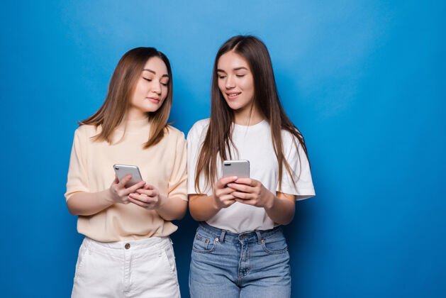 可爱两个多民族的夏季妇女穿着衣服 在蓝色的墙壁上用手机表达兴奋或惊讶年轻爱女人