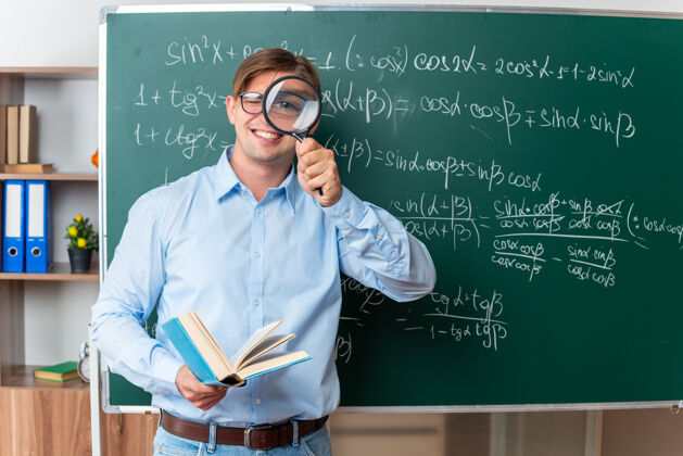 玻璃年轻的男老师戴着眼镜 拿着放大镜和书 兴高采烈地在教室里用数学公式站在黑板旁边讲解功课年轻快乐公式