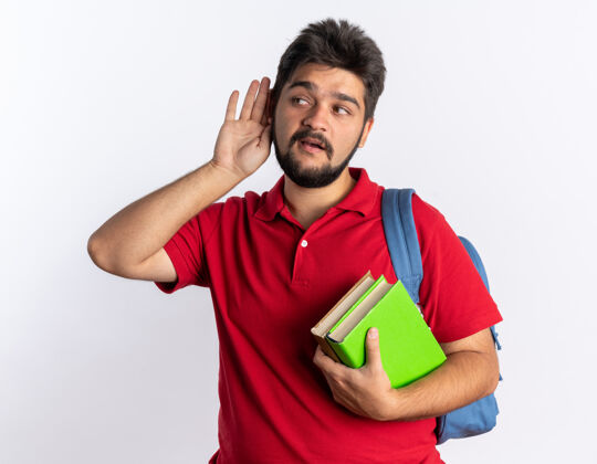 耳朵一个留着胡子的年轻学生 穿着红色马球衫 背着背包 手里拿着笔记本 手捂着耳朵 站在那里听着 看上去很好奇年轻家伙拿着