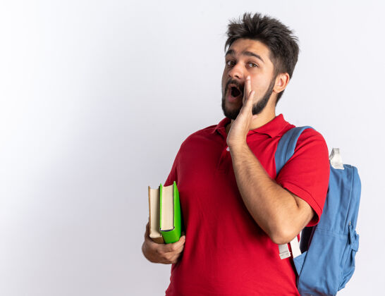 马球年轻的留着胡子的学生 穿着红色马球衫 背着背包 手里拿着笔记本 站着叫着一个手放在嘴上的人打电话笔记本学生