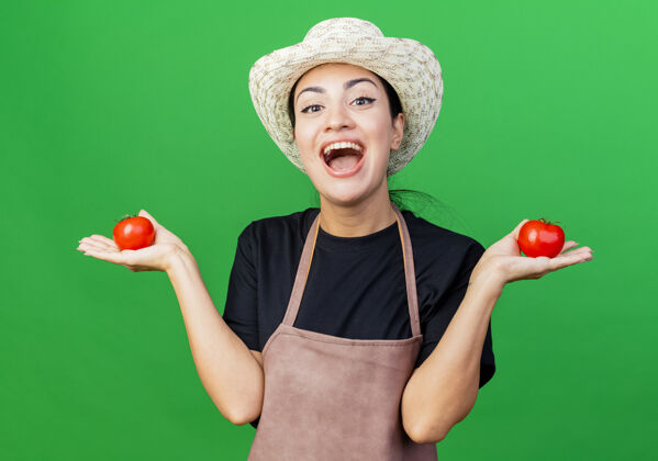 年轻年轻漂亮的女园丁 围着围裙 戴着帽子 手里拿着西红柿 站在绿色的背景上 脸上洋溢着幸福的笑容漂亮抱着围裙