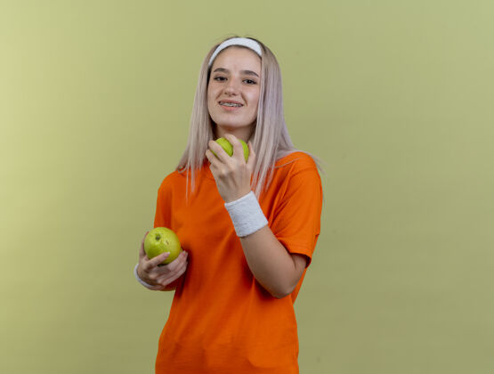 绿色微笑的年轻白人运动女孩戴着牙套戴着头带和腕带拿着苹果女孩橄榄太空
