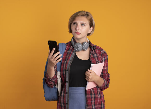 橙色体贴的年轻斯拉夫女学生 戴着耳机 背着背包 拿着笔记本和手机 看着身边学生空间耳机