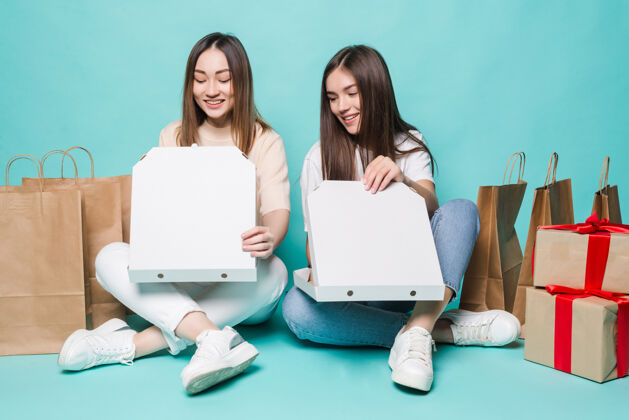 模型微笑着 两个女孩坐在地板上购物袋 在绿松石墙上赠送开放比萨饼友谊多种族支持