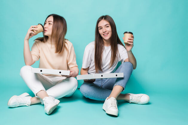女孩两个女朋友坐在地板上喝咖啡去吃披萨隔离在绿松石墙上多民族手模型