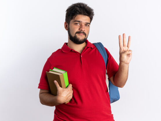 握着年轻的留着胡子的学生 穿着红色马球衫 背着背包 拿着笔记本 面带微笑 三号手指快乐而积极地站着站着正面笔记本