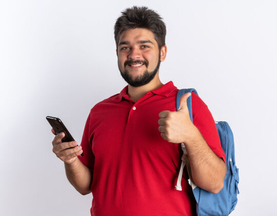 拇指年轻的留着胡子的学生 穿着红色马球衫 背着背包 拿着智能手机 面带微笑 站着竖起大拇指红色年轻马球