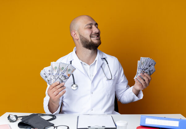 工作快乐的年轻秃头男医生穿着医用长袍和听诊器坐在办公桌旁坐着医生男性