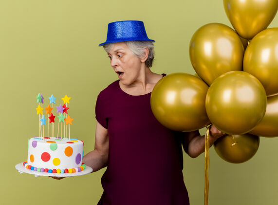 女人惊讶的老妇人戴着派对帽站在氦气球上看着橄榄绿的生日蛋糕站穿氦