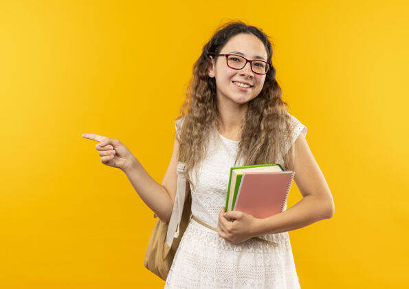 壁板面带微笑的年轻漂亮女生戴着眼镜 背着书包 拿着笔记本 指着隔离在黄色墙上的一侧眼镜人纸条