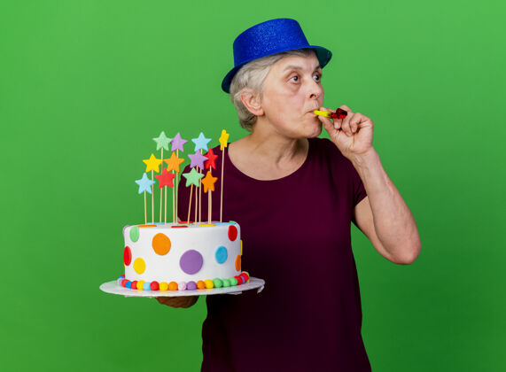 帽子惊讶的戴着派对帽的老太太拿着生日蛋糕吹着口哨望着绿边惊喜女人哨子