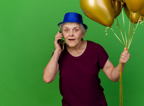 气球惊讶的戴着派对帽的老太太拿着氦气球在绿色的草坪上打电话派对老人女人