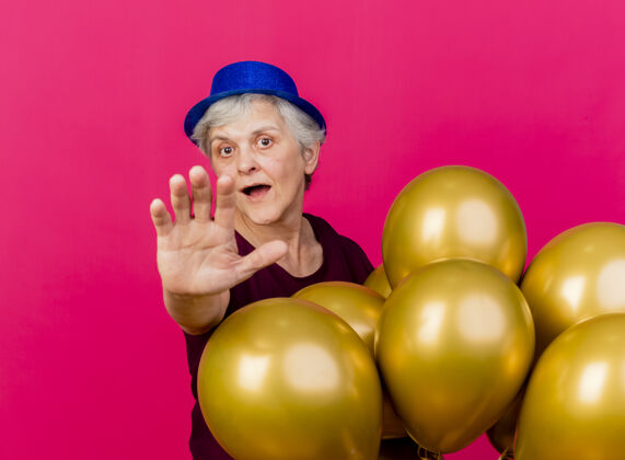 粉色惊讶的老妇人戴着派对帽站着 手里拿着氦气球 伸出粉红色的手穿气球惊喜