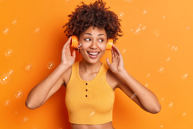 明亮相当满意的卷发女人戴着立体声耳机在耳朵上微笑着广泛地享受完美的声音集中起来穿着不规则的上衣在橙色的工作室墙上与飞行肥皂泡隔离卷发爱人类