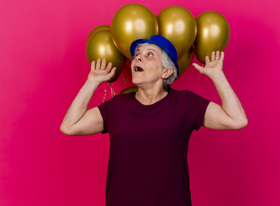 氦一位戴着派对帽的惊讶的老妇人站在氦气球前 举手看着粉红色的气球站起来老人升起