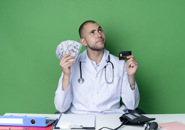 工作体贴的年轻男性医生穿着医用长袍 听诊器坐在办公桌旁 手里拿着工作工具 手里拿着钱和信用卡 孤零零地看着绿色的墙上感情绿色医疗