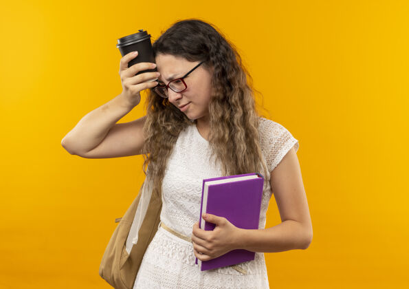 杯子体贴的年轻漂亮的女学生戴着眼镜 背着书包 看着侧碰的头 黄色的墙上隔着塑料咖啡杯黄色年轻表情