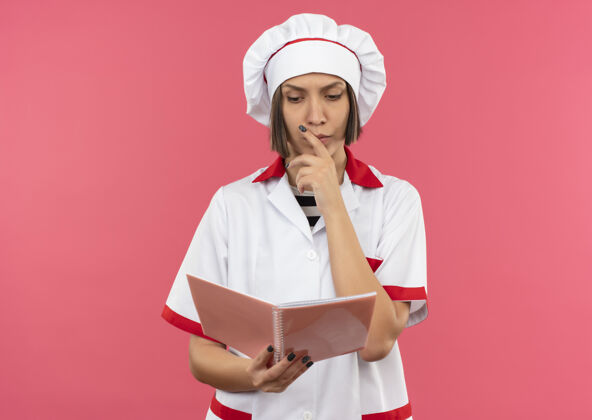 年轻体贴的年轻女厨师穿着厨师制服 拿着便签簿看着 把手放在隔离在粉红色墙上的下巴上姿势笔记脸