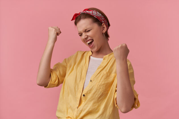 表情身着黄色衬衫 头戴头巾的快乐兴奋的年轻女子 展示赢家的姿态 庆祝战胜粉色墙壁兴奋快乐欢呼