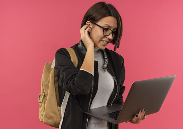 背微笑的年轻女学生戴着眼镜 背着书包 拿着笔记本电脑 把手放在离耳朵很近的粉红色墙上衣服眼镜年轻