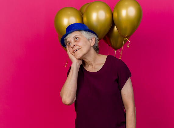 请高兴的老妇人戴着聚会帽站在氦气球前把手放在粉红色的脸上气球前面手