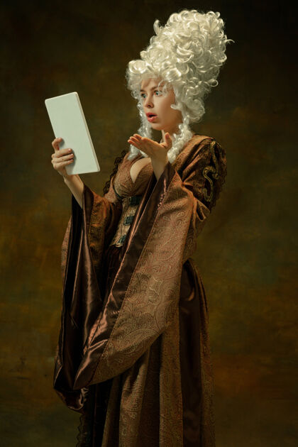 中世纪令人惊讶的使用平板电脑在黑暗的背景下中世纪年轻女子穿着棕色复古服装的肖像作为公爵夫人 王室人士的女性模型时代 现代 时尚 美丽的比较概念公平肖像人