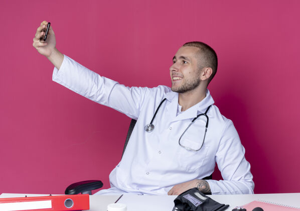 年轻面带微笑的年轻男医生穿着医用长袍和听诊器坐在办公桌旁 带着工作工具 独自在粉红色的墙上自拍长袍感觉拿着