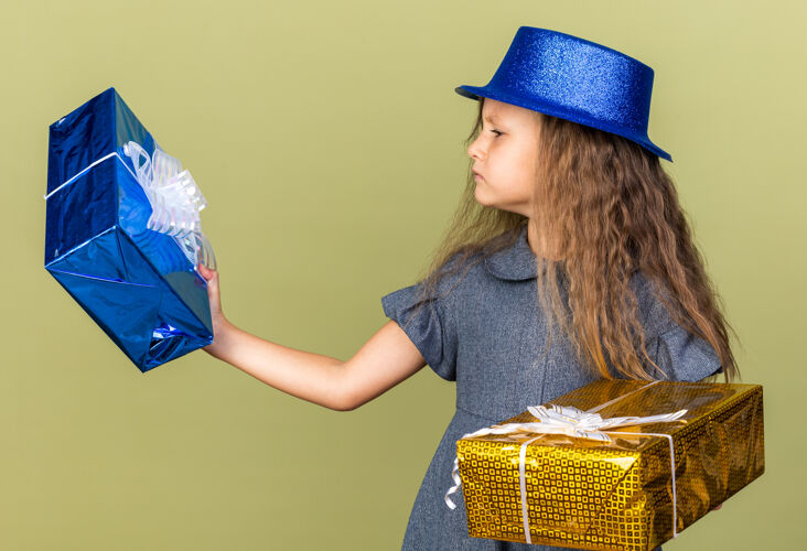 女孩笨手笨脚的金发小女孩拿着蓝色派对帽 看着橄榄绿墙上的礼品盒 还有复印空间无知孤立抱着