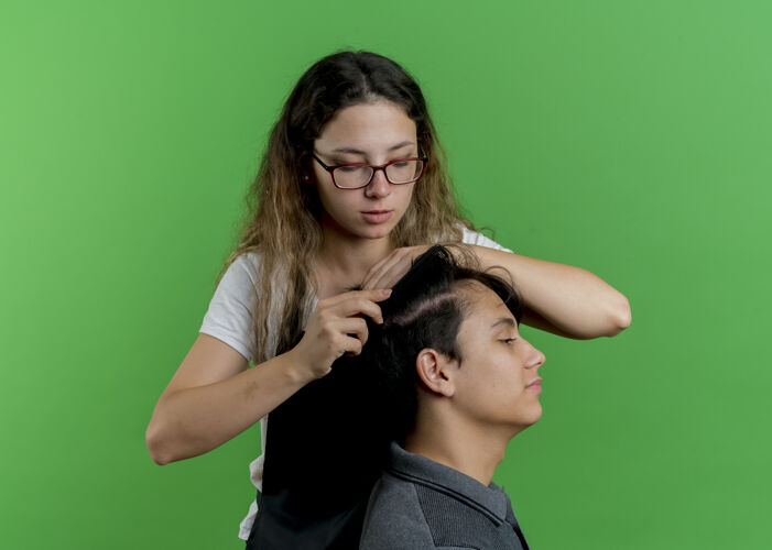 人年轻的专业美发师 围裙里的女人 站在绿墙上梳男顾客的头发脸姿势梳子