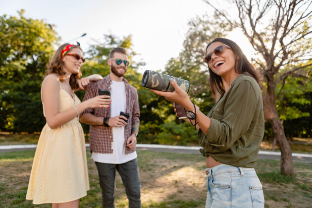 男人年轻时髦的朋友们一起在公园里玩得开心 微笑着用无线扬声器听音乐 夏日风格的季节年轻女性时尚